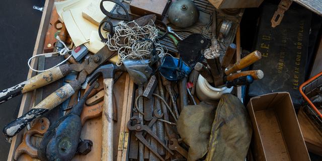 The camera is looking down on a pile of rusty old tools in a wooden tool box: large garden shears, rusty files and pliers, a ball of twine, yellowing paper.
