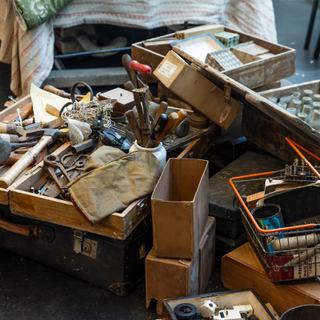 Cluttered boxes cover the floor in a rehearsal room. Including: a scuffed wooden toolbox filled with rusty pliers, hammers and chisels; a crate filled with dirty glass bottles; a shopping basket with paint cans and rollers.