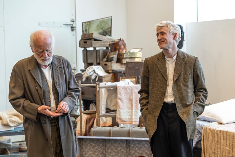 Two men stand in a rehearsal room, in front of a messy pile of furniture and boxes. On the left is an older white-haired man wearing a long brown jacket and white shirt. He is looking down at his hands as he fiddles with a small object. On the left a younger blond man stands with his hands in the pockets of an oversized brown chequered blazer. He is looking towards the other man, cocking his head and smiling.