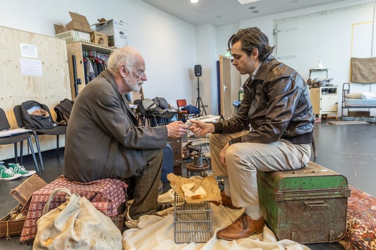 Two actors sit facing each other in a rehearsal room. On the left, an older man in a brown coat and trousers sits on a hamper covered with a pink chequered blanket. On the right a younger man in a leather jacket sits on a rusted green metal chest. The older man looks the younger man in the face, and holds a small object in his hands. The younger man's eyes are down, and he rests his thumb on the small object. Between them is a half eaten sandwich in brown paper.