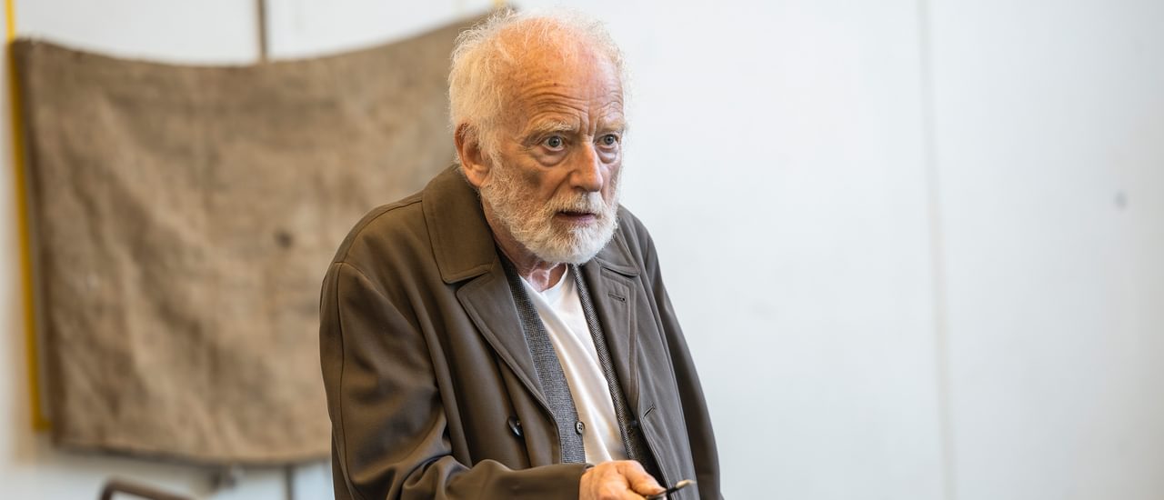 Ian McDiarmid stands in a rehearsal room, with a serious expression on his face. He has wispy white hair and a beard, wears a long brown coat and holds a key in his hand. He is pointing the key out in front of him, as if it might be a weapon.