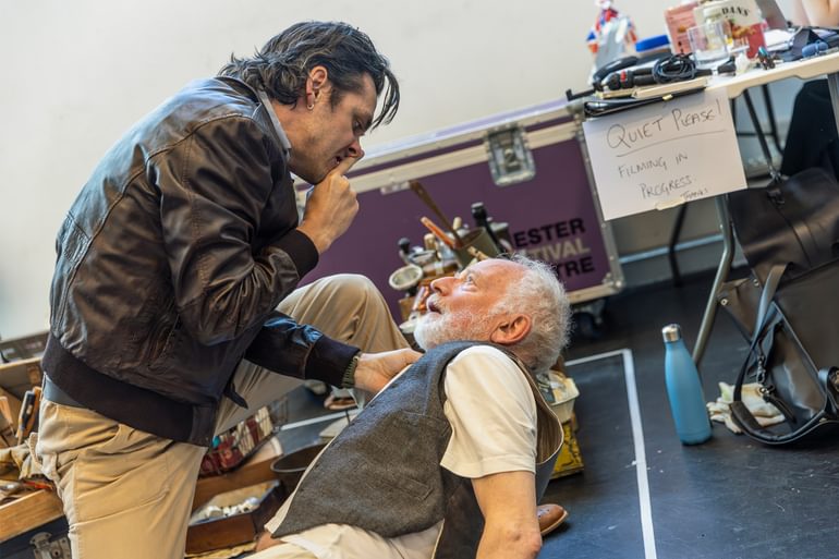 An actor with slicked back dark hair in a brown leather jacket holds a finger to his lips as lunges over an older man aggressively. He holds the older man up off the floor with one hand on his waistcoat collar. In the background are piles of painting and DIY supplies.