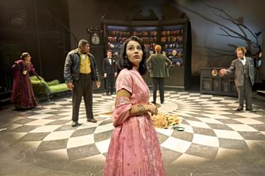 A group of six actors in a theatrical scene stand in varied dramatic poses. They are positioned in front of a dark wooden bookshelf filled with books and decorative items, underfoot is a patterned floor design. At the front of the image, a woman in a pink embroidered dress looks out with a concerned expression.