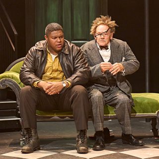 Two men sit on a green chaise lounge. The man on the left wears a brown leather bomber jacket and mustard yellow shirt, he looks concerned. The man on the right is in a three-piece grey tweed suit and bowtie, he looks shaken up with his glasses askew.