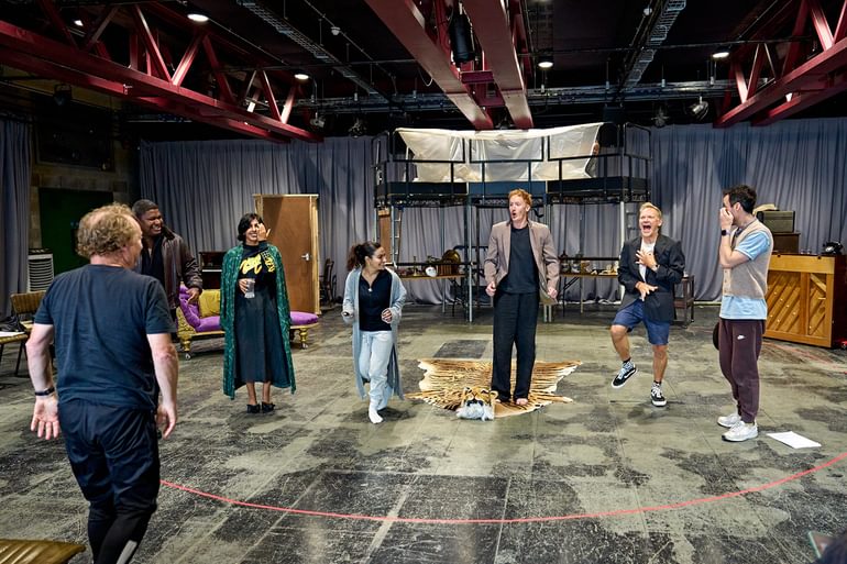 A group of people stand in a line in a rehearsal room, laughing, moving and gesturing. On the floor is a fake tiger rug.