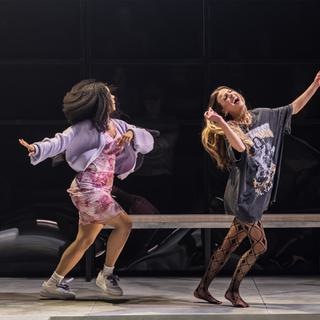Two young women dance around on stage, laughing and smiling. The woman on the left is wearing white trainers, a pink mini dress and a lilac cardigan. Her dark curly hair and the cardigan are flying out as she moves forward, with her arm stretched out behind her. The woman on the right has her eyes closed and her head up in laughter as she dances or runs forward. She is wearing a baggy graphical t-shirt, fishnet tights with black triangles on them, and has long blonde hair flowing behind her.