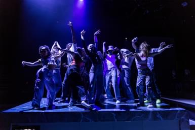 A group of young people dance on top of a kitchen island on a stage. They are stood together, with their arms raised and twisted, mid-dance move. They are wearing party clothes - trainers, jeans, crop tops and bucket hats - and are lit with dramatic blue lighting.