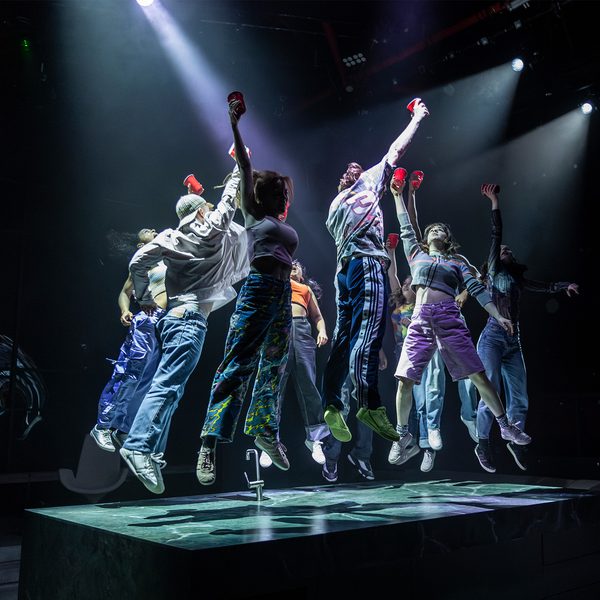 A group of young people dance on top of a kitchen island on a stage. They are all jumping in unison, with their feet off the ground and an arm stretched above their head holding a plastic cup. They are wearing colourful, sporty clothes - trainers, jogging bottoms and shorts. They are lit with dramatic white spot lights.