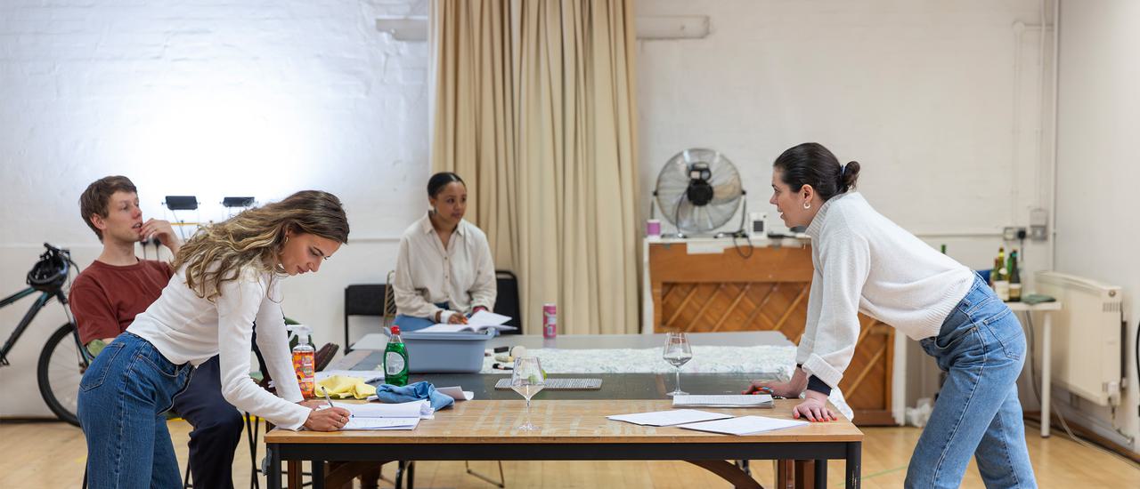 Four people are dotted around a large tabe in a rehearsal room. On the left is a blonde young woman in blue jeans and a white shirt. She leans onto the table, writing in a script. Behind her, a man in a red t-shirt leans back on his chair. In the background, a woman with dark hair in a white shirt sits on a chair, looking toward a woman on the right who is also leaning on the table.