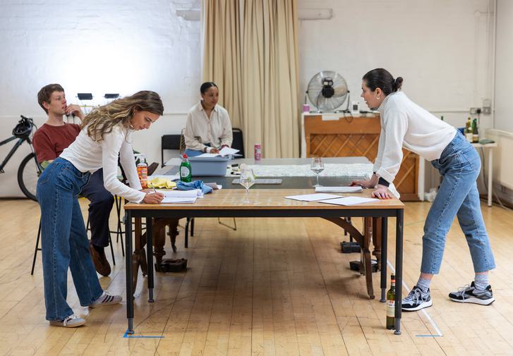 Four people are dotted around a large tabe in a rehearsal room. On the left is a blonde young woman in blue jeans and a white shirt. She leans onto the table, writing in a script. Behind her, a man in a red t-shirt leans back on his chair. In the background, a woman with dark hair in a white shirt sits on a chair, looking toward a woman on the right who is also leaning on the table.