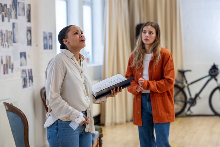 A young woman in a white shirt and blue jeans holds a notebook in one hand and looks up worriedly. Behind her, a young woman with long blonde hair wearing an orange jacket and blue jeans looks on with a concerned expression.
