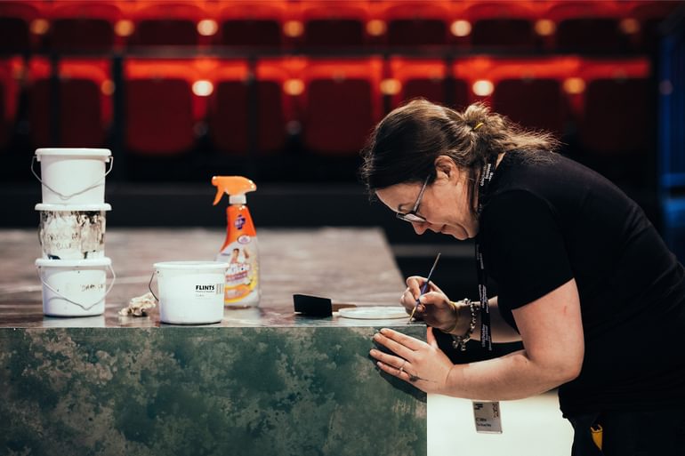 A member of the crew is photographed side on, carefully painging a piece of scenery with a small paint brush. In front of her, on top of the scenery, are four white buckets of paint and a cleaning product in a spray bottle. Out of focus in the background the red theatre seats can be seen.