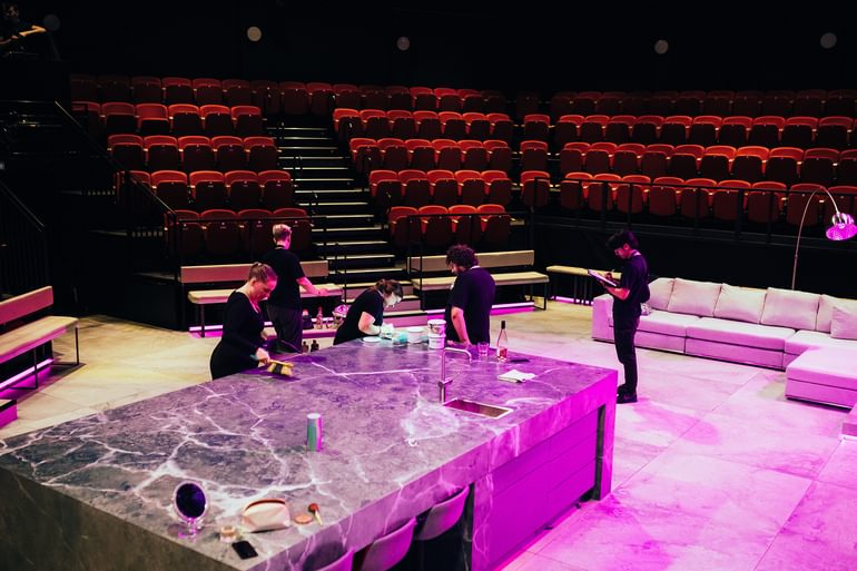The Minerva Theatre stage photographed from above. There is a large kitchen island with a marble effect, with three bar stools tucked underneath it. On top of the island there is a sink with a silver tap, a makeup bag with a mirror and makeup items, two phones, a tea towel, a bottle of wine and a glass. Five members of the show crew are gathered around. They are all very busy and concentrating carefully on their different tasks. One checks a clip board, one cleans the kitchen island with a brush, one shines a torch on the island where another is painting. Behind them there is a large, grey sofa with a silver lamp over it. The red theatre seats surround the stage, which is lit in pink.