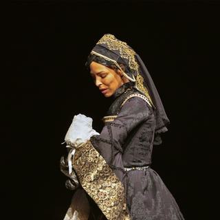 A woman in regal traditional Tudor costume stands side on, holding a baby dressed in white