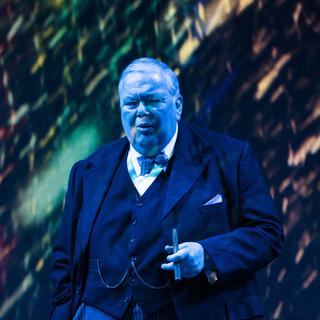 A man resembling Winston Churchill, dressed in a smart black suit and bowtie, stands with a cigar in his hand. He is mid-speech, with a questioning expression as he talks.