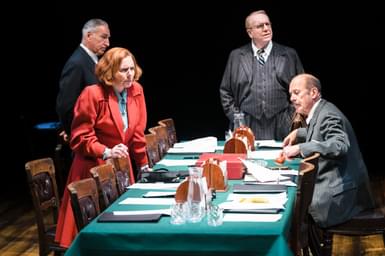 Three men and a woman are gathered around a conference table, looking deep in thought. The table is covered in documents that are spread open. The men all wear dark grey pin-striped suits, while the woman wears a bright red coat.