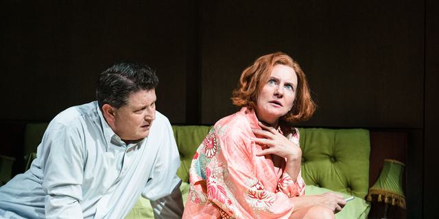 A man and woman sit together on a green double bed in a state of undress. She wears a pink silk robe, smoking, while he wears only his shirt. He is leaning into her inquisitively as she looks off into the distance with a quizzical expression.