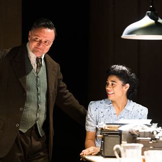 A woman sits at the table in front of a typewriter, lit from above by a ceiling lamp, while a man is standing next to her. He is casually resting one hand on his hip and the other on the back of her chair. They are both smartly dressed in a 1940s style. They have light, joyful expressions.
