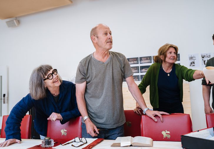 Three performers stand next to a long table that is surrounded with red leather chairs. The lady on the left wears a dark blue shirt and thick framed glasses. She leans over the table and smiles off to the side. The man next to her is looking in the same direction, talking, with one hand resting on the back of a chair. He wears a grey t-shirt and dark leather jeans. The woman on the right is leaning forward, passing a piece of paper to someone out of shot. She wears a grey cardigan and has her medium brown hair tied up. She is smiling and looking up. They all look relaxed and happy.