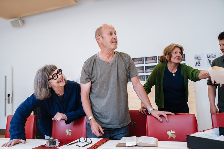 Three performers stand next to a long table that is surrounded with red leather chairs. The lady on the left wears a dark blue shirt and thick framed glasses. She leans over the table and smiles off to the side. The man next to her is looking in the same direction, talking, with one hand resting on the back of a chair. He wears a grey t-shirt and dark leather jeans. The woman on the right is leaning forward, passing a piece of paper to someone out of shot. She wears a grey cardigan and has her medium brown hair tied up. She is smiling and looking up. They all look relaxed and happy.