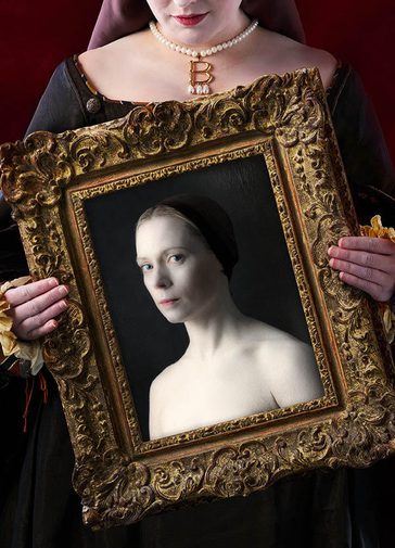 A woman's hands can be seen holding an ornate golden picture frame. The bottom of the woman's face is visible above the frame, she has a slight smile, red lipstick and is wearing a pearl neckace with a golden 'B' on it. Inside the picture frame is an image of a serious looking woman. The top of her chest and shoulders are exposed, and she wears a brown cap with her hair pulled back.