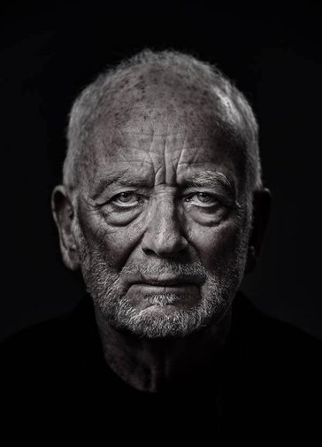 An elderly man (Ian McDiarmid) stares at the camera. His face is wrinkled and looks stern and dramatic, with shadows under his eyes and chin. He is wearing a black shirt, and his face in lit in a silvery colour.