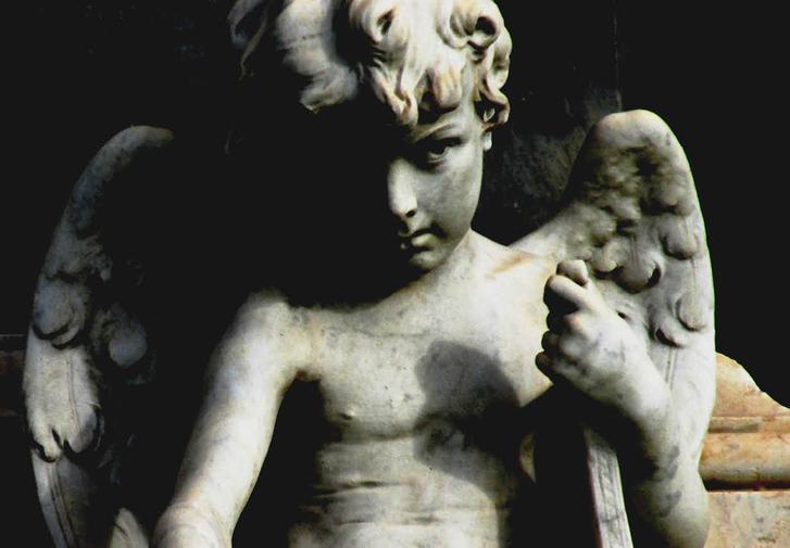 A statue of a curly haired cherub, holding some papers in his hand. The statue is in shadows, with the left side of the face and half its wing in darkness. It looks dramatic.