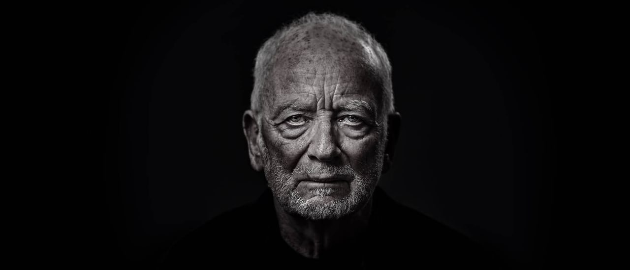 An elderly man (Ian McDiarmid) stares at the camera. His face is wrinkled and looks stern and dramatic, with shadows under his eyes and chin. He is wearing a black shirt, and his face in lit in a silvery colour.