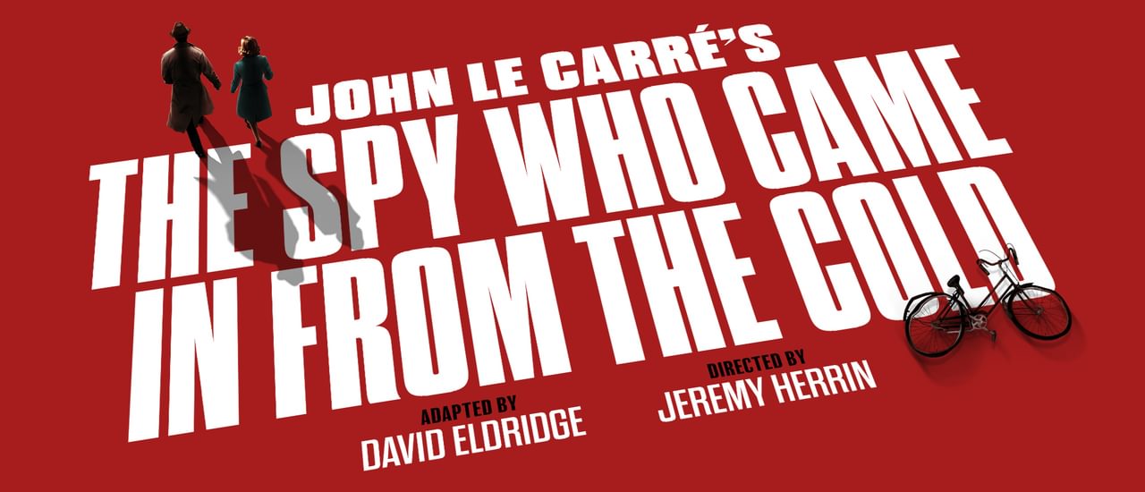 A red graphic with bold, white all-caps text that reads 'John le Carre's The Spy Who Came In From The Cold, adapted by David Eldridge, directed by Jeremy Herrin'. There is a small graphic of a couple, the man wearing a spy-style trench coat and hat, and a bike.