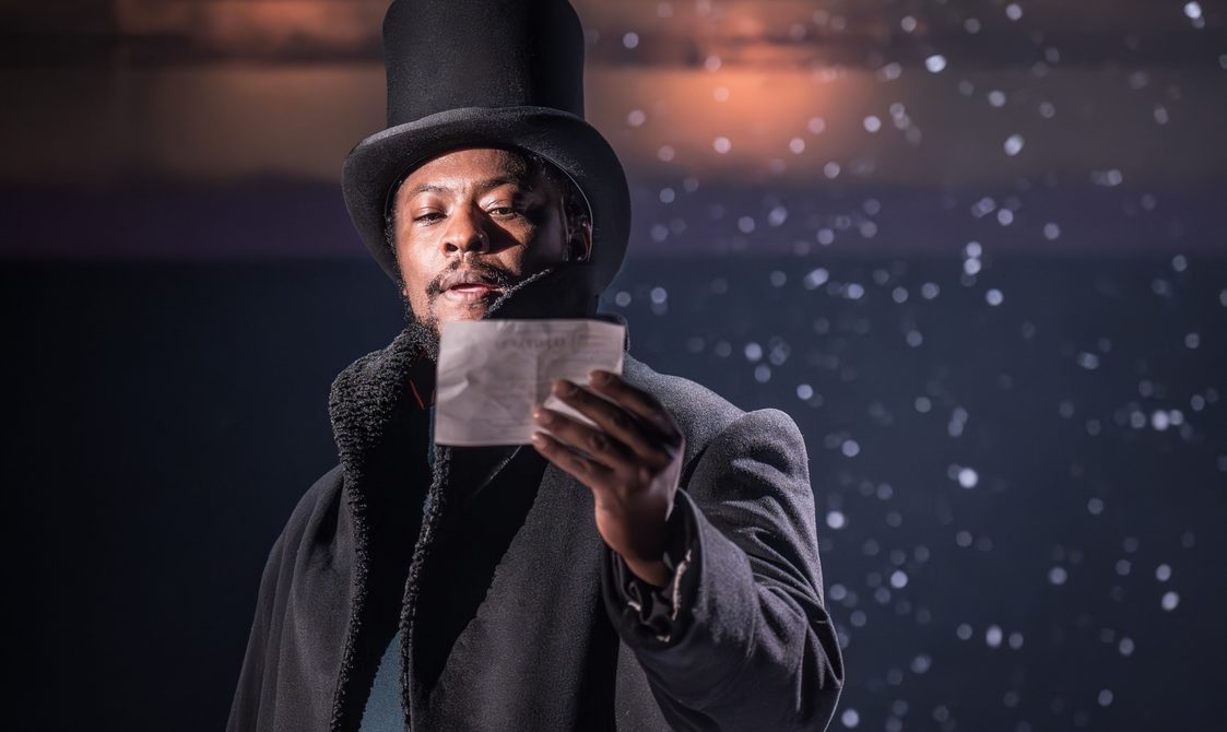 A man with a black top hat and a black overcoat is reading from a piece of paper he is holding. There is snow falling delicately behind him.