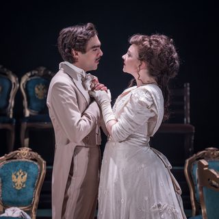 A man and a woman are stood looking at each other longingly, holding hands together are resting them on his chest. They are both dressed exquisitely; he has on an elegant taupe suit and she is wearing a cream gown with ruched sleeves and a silk cinching belt.