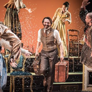 A jovial scene with several performers all in neutral-toned Victorian era costumes smiling and dancing. There is white confetti floating all around the stage. In the centre, a man with dark hair and facial hair is smiling brightly. He is holding a vintage leather suitcase in each hand.
