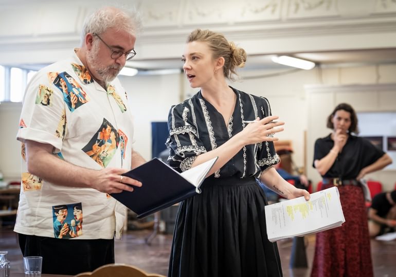 A man with white hair and a beard is reading a notebook which he is holding. He is wearing glasses and a white shirt with various colourful portraits. Next to him, there is a woman with blonde hair talking to him. She is wearing a black blouse with white detailing and a black skirt. She is holding a script with a lot of text highlighted in one hand and a pen in the other. In the background, there is a woman wearing a black top and long red skirt looking at them with one hand on her hip and the other resting on her chin, as if in thought.