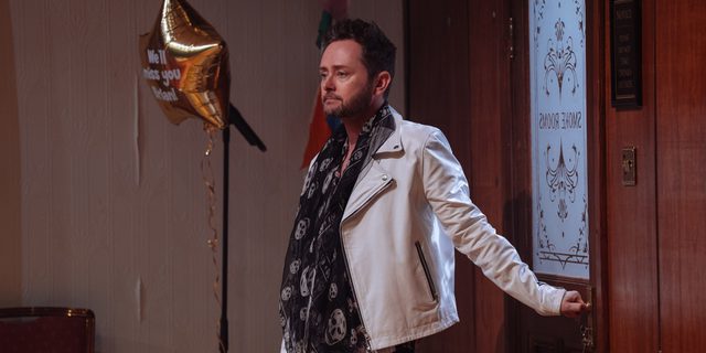 A bearded man with a slightly glum expression wearing a white leather jacket and a black scarf with grey skulls is stood with his hand grasping the handle of a door which reads 'Smoke Rooms'. Next to him is a gold foil star balloon which reads 'We'll miss you Brian!'