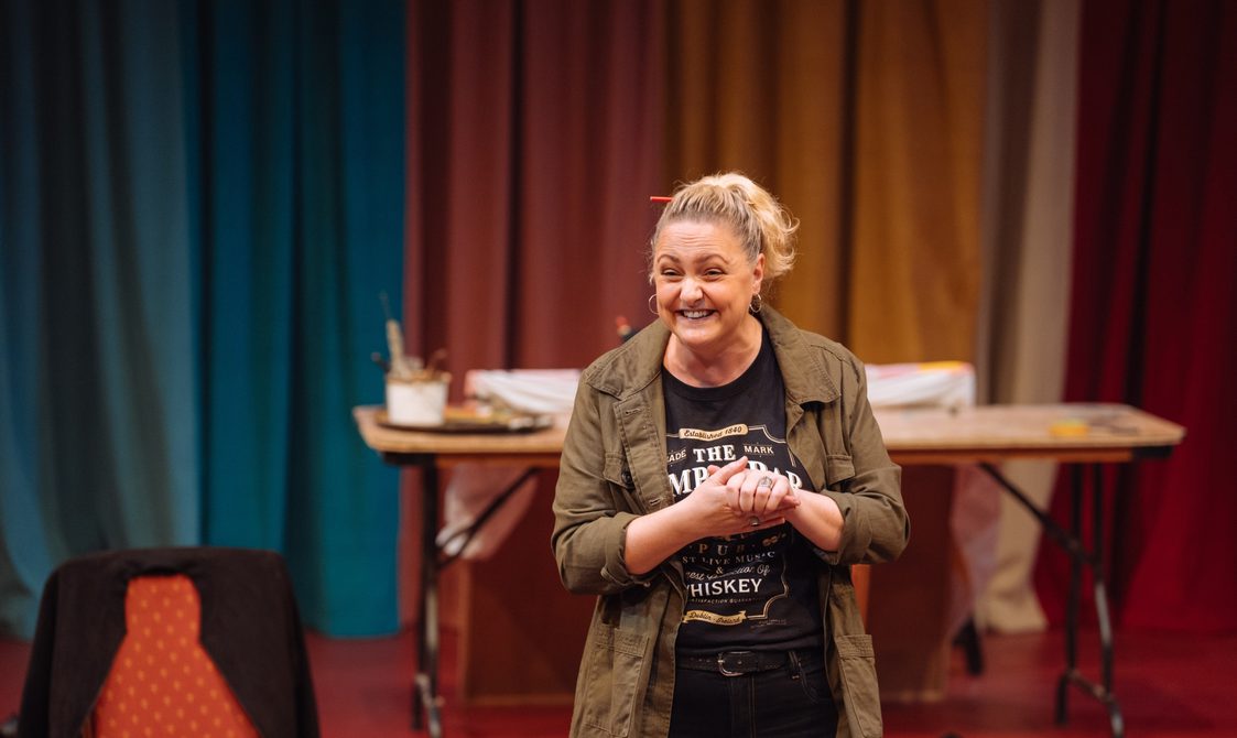 A woman with her blonde hair in a ponytail with an orange pencil tucked through it is smiling excitedly. Her hands are clasped in front of her. She is wearing a casual khaki jacket over a black graphic t-shirt. In the background, there is a table with art supplies in front of swathes of colourful curtains.