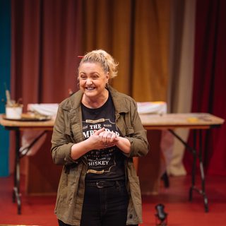 A woman with her blonde hair in a ponytail with an orange pencil tucked through it is smiling excitedly. Her hands are clasped in front of her. She is wearing a casual khaki jacket over a black graphic t-shirt. In the background, there is a table with art supplies in front of swathes of colourful curtains.