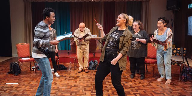 A group of performers are stood in a semi-circle, singing gloriously while reading from folders. In the centre, a woman with blonde hair is using a pencil like a conductor's baton. They are in a dated rehearsal room, with retro wooden floors, colourful curtains and fairy lights.