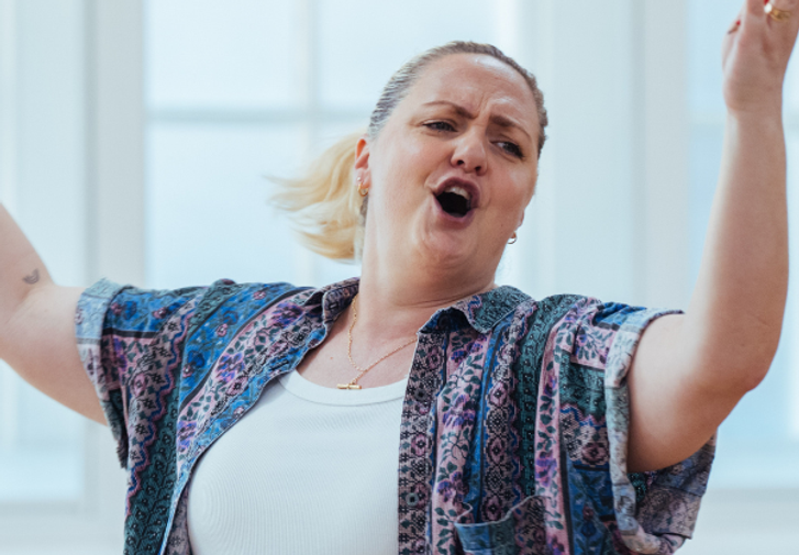 A woman with blonde hair in a ponytail has her arms raised above her head and is singing passionately. She is wearing a casual, colourful shirt over a white vest.