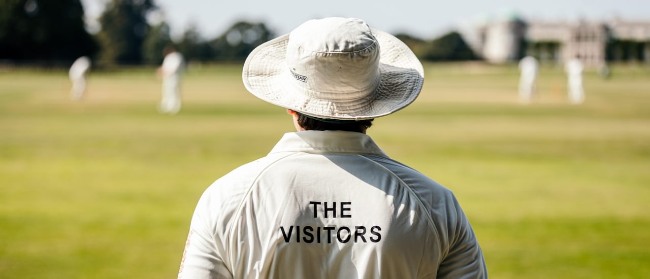 The back of a cricketer in whites with a hat on overlooking a green with cricketers in the distance. On the back of his shirt reads The Visitors in black type.