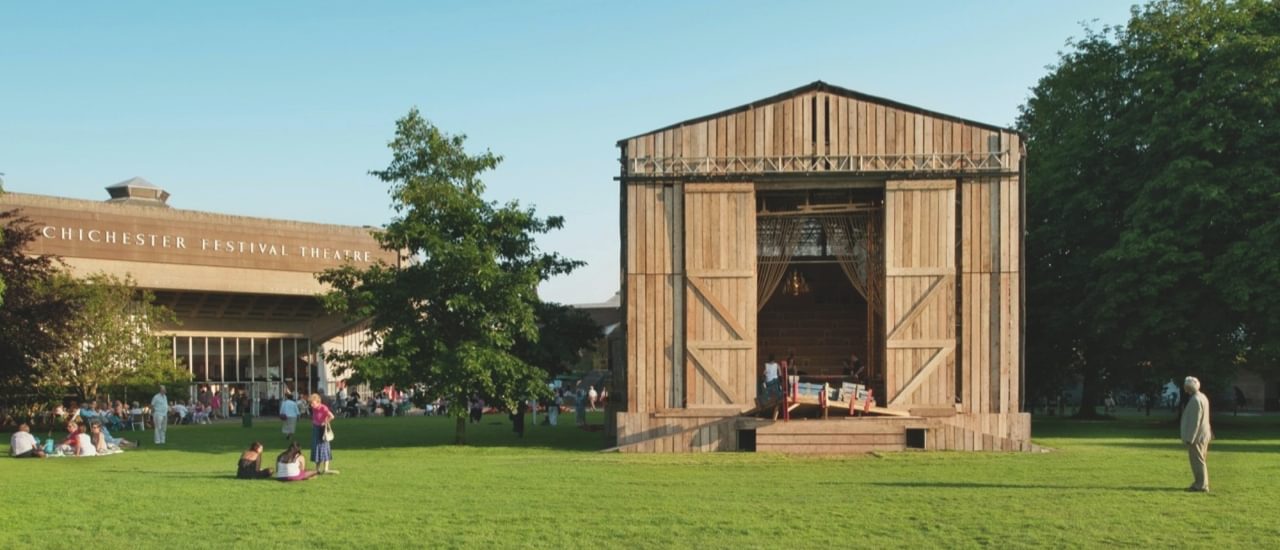 A temporary wooden stage structure is stood next to the Festival Theatre. The structure has a classic barn shape. The sky is very blue and the trees are a vibrant green.