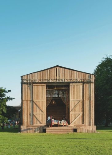 A temporary wooden stage structure is stood next to the Festival Theatre. The structure has a classic barn shape. The sky is very blue and the trees are a vibrant green.