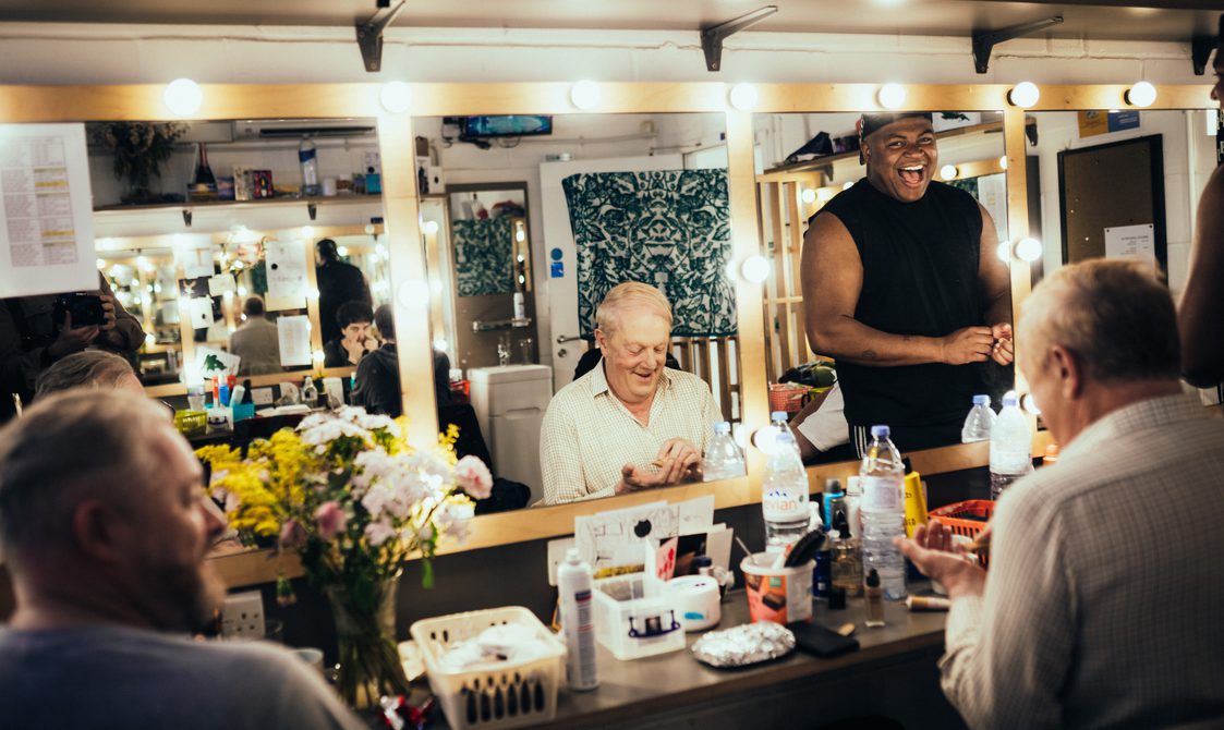 Inside a dressing room, two actors are seated and getting ready, a third is standing smiling brightly at the mirror.