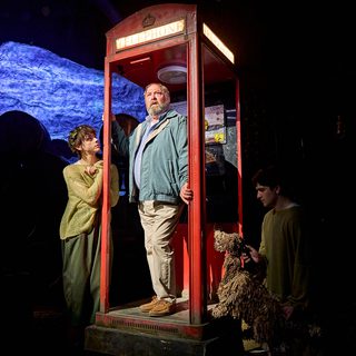 An older man wearing casual clothes is stood inside a worn-down red telephone box. His hands are clasping the doorframe, and he is looking slightly worried. To his right, a young man dressed in an almost threadbare green jumper with a crown of leaves in his hair is gazing up at him inquisitively. On the other side, a shaggy dog puppet operated by a puppeteer is also looking at the man in the phone box.
