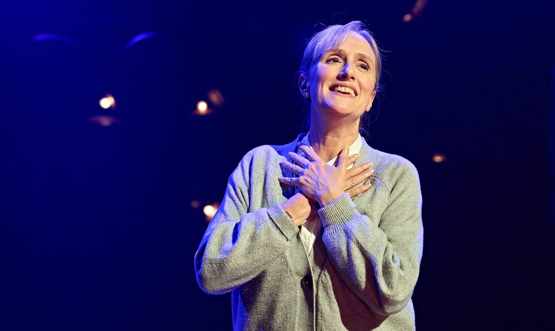 A woman wearing a grey cardigan has her hands protectively placed over one another in the middle of her chest. She has a bright smile on her face as she looks off into the distance.