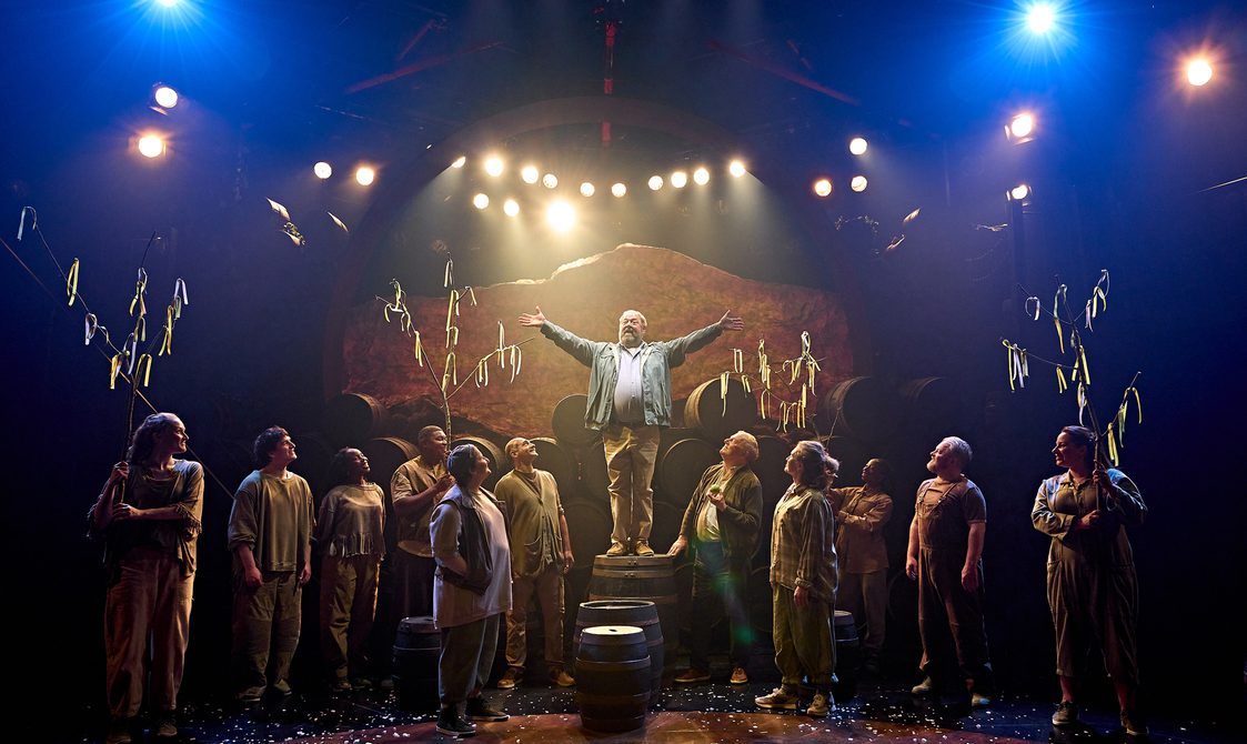 Many characters are gazing wondrously at a man is standing atop a barrel in the centre of the stage. He has his arms wide open. Some of the ensemble are holding tree branches with ribbons tied to them. There are many barrels stacked in the background and others placed amongst the group of people.