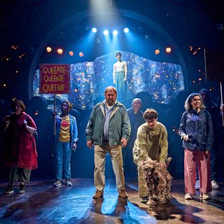 An eclectic mix of characters are standing, with an older man in the centre, looking tired. There is a vicar, a woman holding a microphone, a puppeteer operating a shaggy dog puppet, another woman wearing a garish magenta poncho and another holding a pink placard which has ‘Queenie’ written three times in bright yellow capital letters. Behind this group is a young man standing at the top of a stack of barrels looking down at them, with twinkling lights in the background.