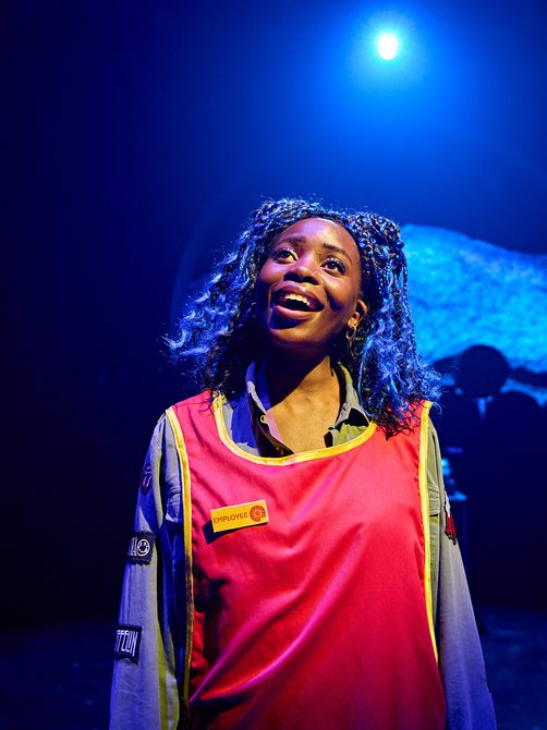 A young woman is singing with a gleeful smile. She is dressed in a work uniform; a red tabard with bright yellow trim with a badge that reads ‘employee’ accompanied by a seashell logo. Underneath she has a loose-fitting grey shirt adorned with various patches of rock band logos on the sleeves.