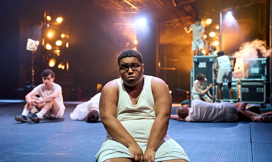 A boy in a white vest and shorts and big, round glasses sits on the edge of the stage. He has a sad expression with his bottom lip protruding. Behind him, several other boys are lying around and others are climbing on flight cases surrounded by haze.