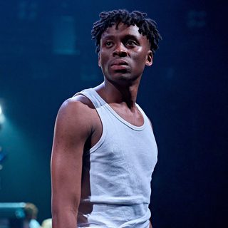 A boy in a white vest looks over his shoulder. He has a neutral expression. The lighting is blue.