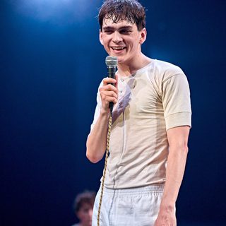 A boy in cream and white undergarments is holding a microphone up to his mouth. His hair is wet from sweat and he is smiling wryly.